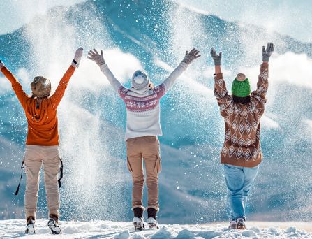 3 Menschen tragen Winterkleidung und werfen pulvrigen Schnee in die Luft. Im Hintergrund sieht man eine schneebedeckte Winterlandschaft