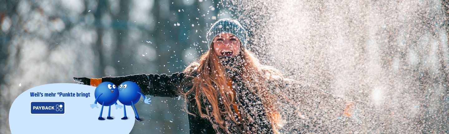 Eine Frau in Winterkleidung wirft pulverartigen Schnee in die Luft und lacht herzlich