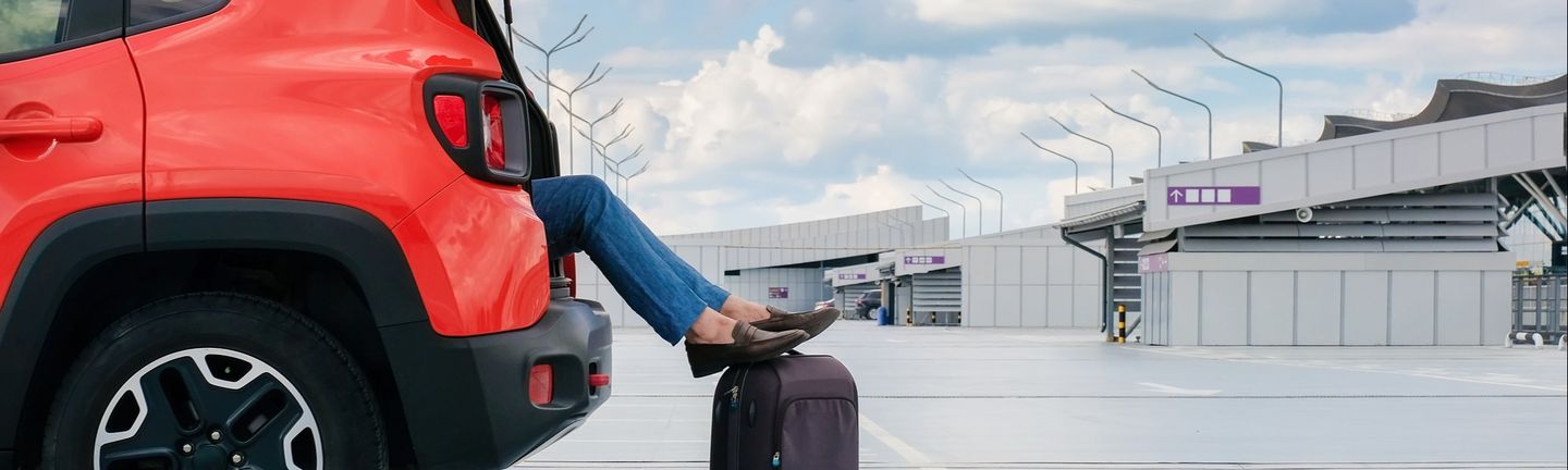 Frauenbeine ragen aus dem Kofferraum eines roten Autos und sind auf einen Koffer gelehnt auf einem Flughafenparkplatz