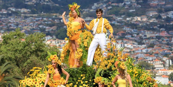 Auf einem, mit Blumen geschmückten Wagen, steht ein komplett gelb gekleidetes Paar, Händchenhaltend und winkt in die Menge. Der Wagen ist mit Blumen geschmückt und im Hintergrund sieht man ein madeirisches Dorf.