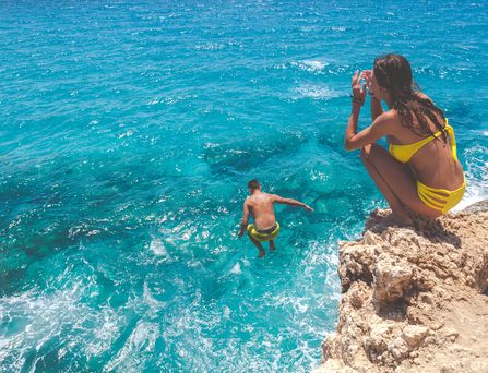Ein Junge springt von einer Klippe ins türkise Meer. Ein Mädchen im gelben Bikini sitzt am Rand der Klippe in der Hocke.