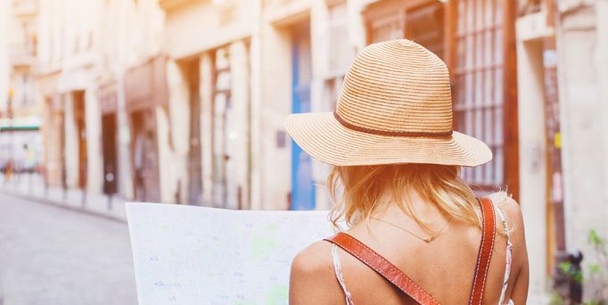 Eine Frau mit Sonnenhut und RUcksack läuft durch eine hübsche Straße und hält eine Landkarte in der Hand, auf die sie blickt.