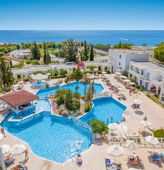 Direkt am Meer liegt eine Hotelanlage mit einer Poollandschaft zwischendrin immer wieder grüne Bäume und viele Sonnenschirme.