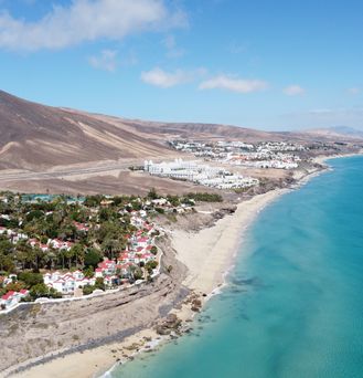 Eine karge, trockene, braune Berglandschaft reicht bis an den hellen, Sandstrand. Einzelne weiße Häusergruppen mit orangenem Dach und grünen Bäumen stehen am Strand.