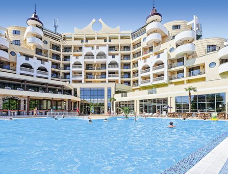 An einer verspiegelten Glaswand tropft ein künstlicher Wasserfall in einen türkisen Wassergraben der am Gebäuderand entlang führt. Daneben eine Wiese mit Blumen und Palmen.
