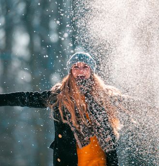 Eine junge blonde Frau mit Mütze und Handschuhen breitet lachend die Arme aus und wirbelt pulvrigen Schnee hoch.