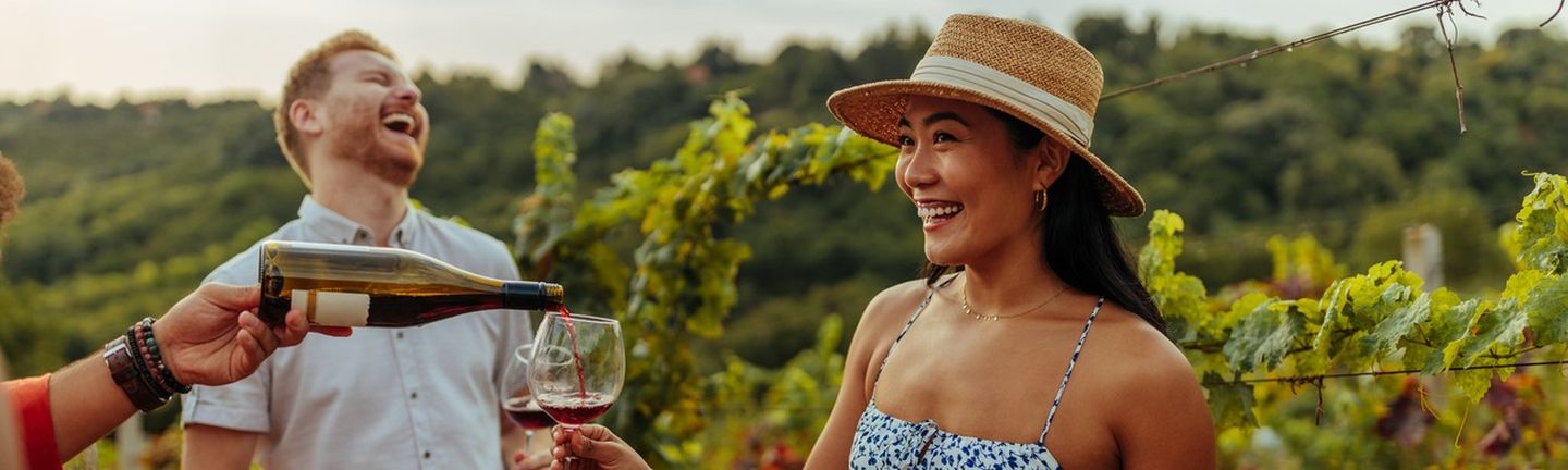 Eine Frau steht im Sommerkleid mit Sonnenhut lachend in einem Weinanbaugebiet. Sie hält ein Weinglas vor sich in welches gerade Rotwein geschüttet wird. Im Hintergrund steht ein Mann, welcher herzhaft lacht, ebenfalls mit einem Rotweinglas in der Hand.