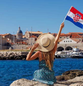 Eine junge Frau mit Sonnenhut und Kleid sitzt auf einem Felsen am Hafen und blickt auf das tiefblaue Wasser und die gegenüberliegende Stadt. Sie hält eine kroatische Flagge in der rechten Hand und hält sie hoch.