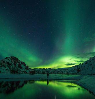 Eine Person steht an einem Fluss, indem sich grüne Polarlichter spiegeln. Im Hintergrund sieht man schneebedeckte Berge.