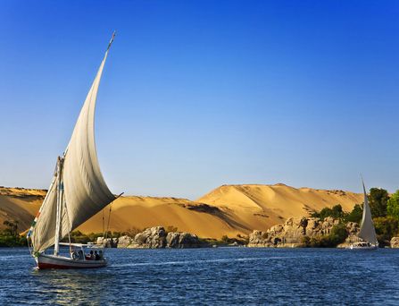 Ein Segelboot auf blauem Wasser, im Hintergrund Sanddünen.