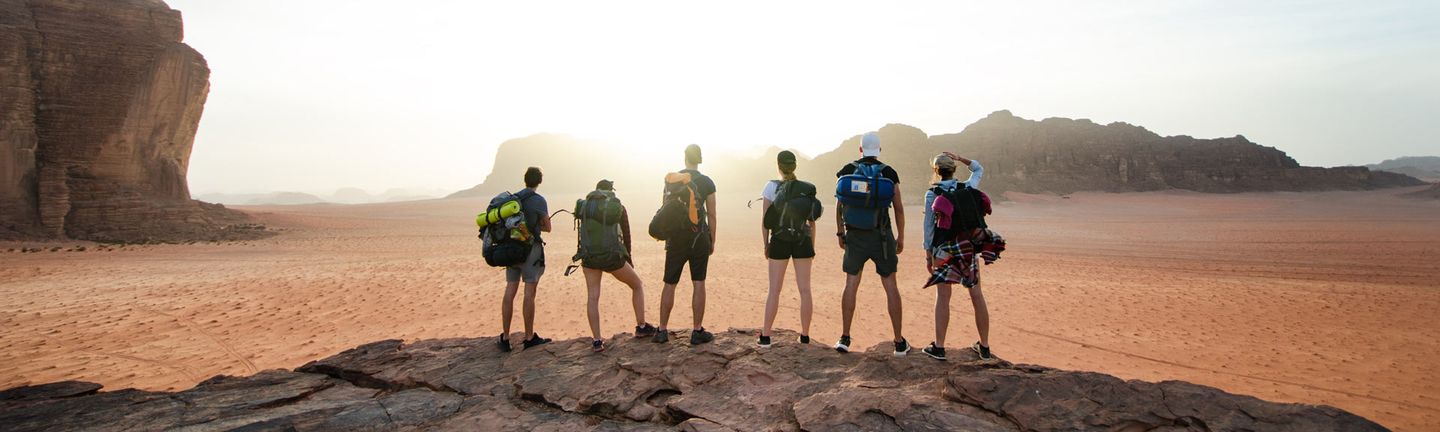 Eine Gruppe aus sechs jungen Menschen mit Rucksäcken und Outdoorausrüstung steht auf einem Felsen und blickt in eine Felslandschaft.