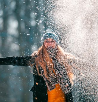 Eine junge blonde Frau mit Mütze und Handschuhen breitet lachend die Arme aus und wirbelt pulvrigen Schnee hoch.