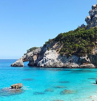 Kristallklares, türkises Wasser an einer steinigen, steilen Bucht. Ein paar Menschen stehen auf einem Felsen.