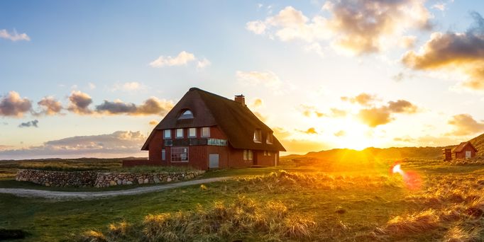 Ein großes rotes Haus mit Strohdach steht einsam in einer weiten, grünen Landschaft. Der Himmel ist von der untergehenden Sonne rosa-gold gefärbt und ganz rechts steht ein rot-weißer Leuchtturm.