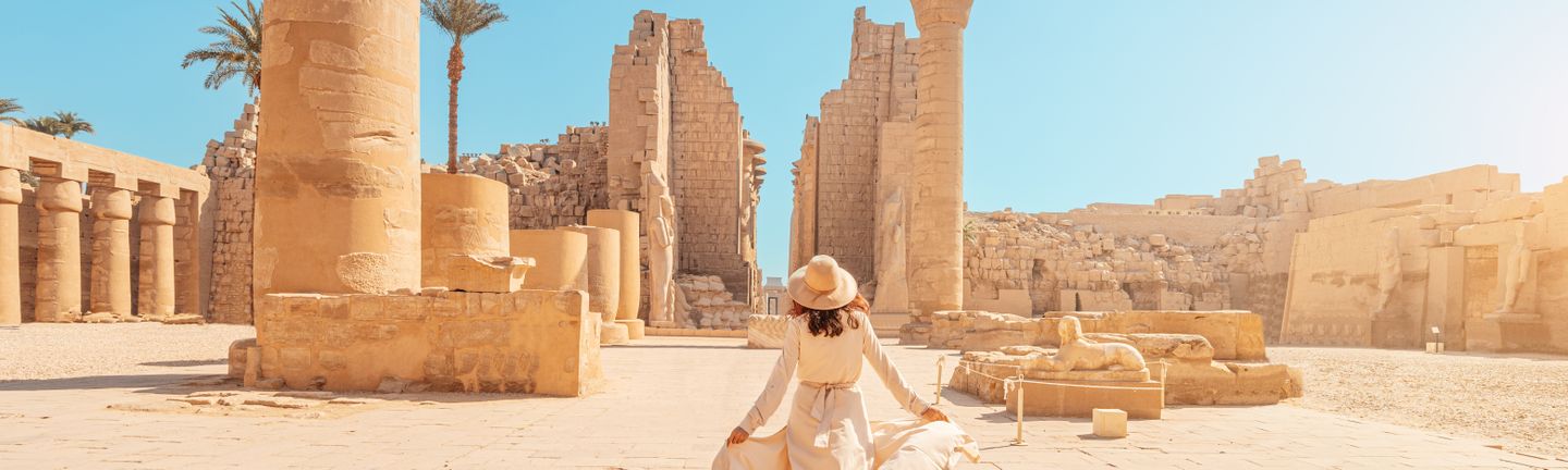 Eine in beige gekleidete Frau mit Sonnenhut läuft durch eine Ruinenlandschaft. Der Himmel ist strahlend blau.