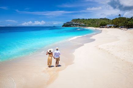 Ein Paar mit Sommerkleidung läuft Hand-in-Hand an einem weißen Sandstrand mit kristallklarem türkisen Wasser entlang.