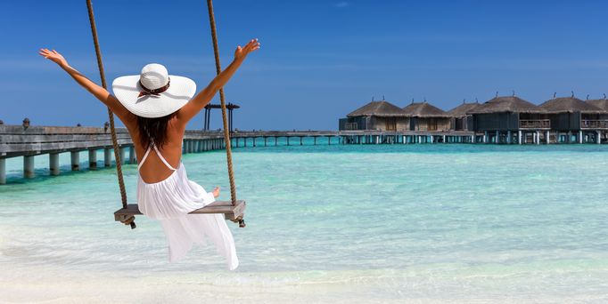 Malediven Urlaub: Eine Frau mit weiße Sommerkleid und Hut sitzt auf einer Schaukel am Strand.