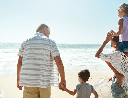 Eine Familie, bestehend aus einer Frau, 2 Kindern im Alter von ca. 4 bis 6 Jahren und einem älteren Mann, laufen an einem Sandstrand aufs Meer zu. Das Mädchen sitzt bei der Frau auf den Schultern und der Junge geht an der Hand des Mannes.