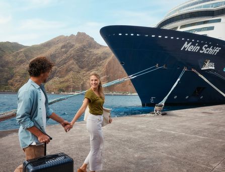 Ein lachendes Paar geht mit Gepäck auf das Kreuzfahrtschiff "Mein Schiff" zu.