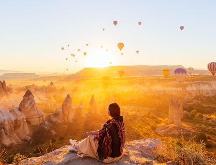 Eine junge Frau sitzt inmitten einer bizarren Berglandschaft und blickt in die Ferne. Die Sonne geht gleich hinter einem Berg unter und taucht die Szene in ein warmgelbes Licht. Der Himmel ist übersäht von unzähligen Heißluftballons.