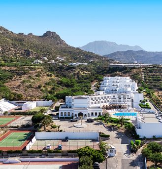 In einer begrünten Berglandschaft befinden sich eine weiße Hotelanlage mit Pool und mehreren Sportplätzen.