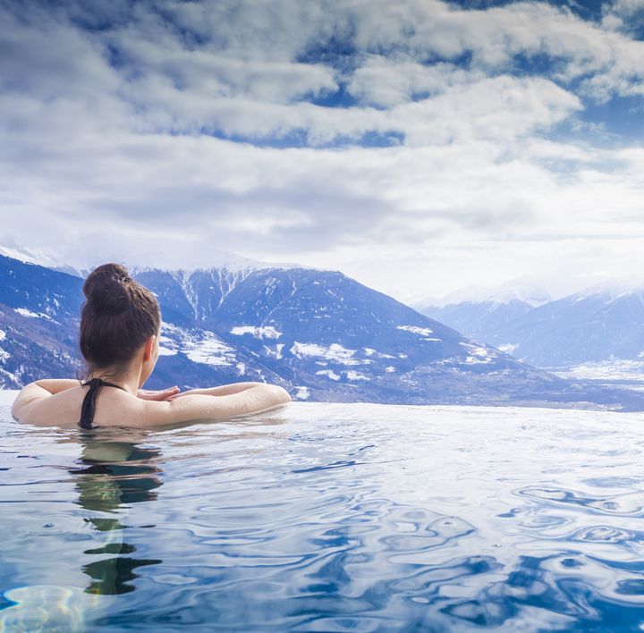 Eine Frau mit braunem Dutt lehnt am Rand eines Infinitypools mit Blick auf die verschneiten Bergspitzen.