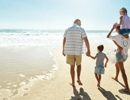 Eine Familie, bestehend aus einer Frau, 2 Kindern im Alter von ca. 4 bis 6 Jahren und einem älteren Mann, laufen an einem Sandstrand aufs Meer zu. Das Mädchen sitzt bei der Frau auf den Schultern und der Junge geht an der Hand des Mannes.