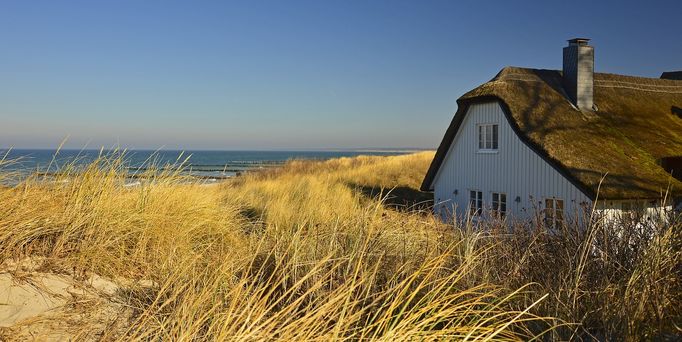 Zwischen Dünen am Meer steht ein weißes Haus mit Strohdach.