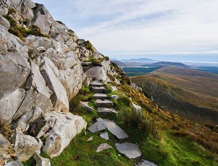 Eine Treppe aus Steinen führt an einer felsigen Mauer hoch. Rechts am Abgrund das Landschaft der Highlands.