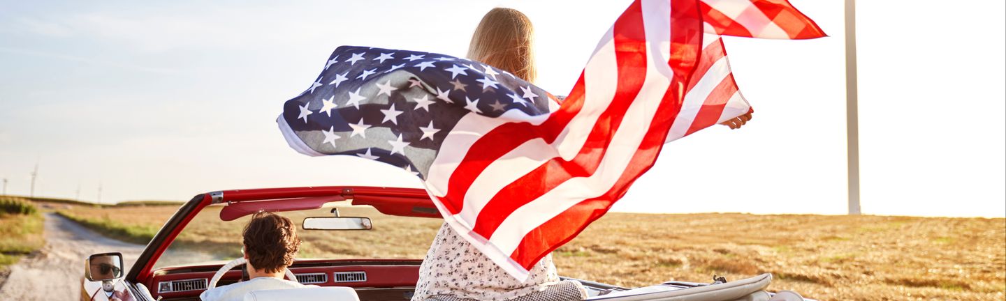 Ein Pärchen fährt in einem Oldtimer Cabrio eine Landstraße entlang und die Beifahrerin schwingt eine USA Flagge im Wind hinter sich mit.