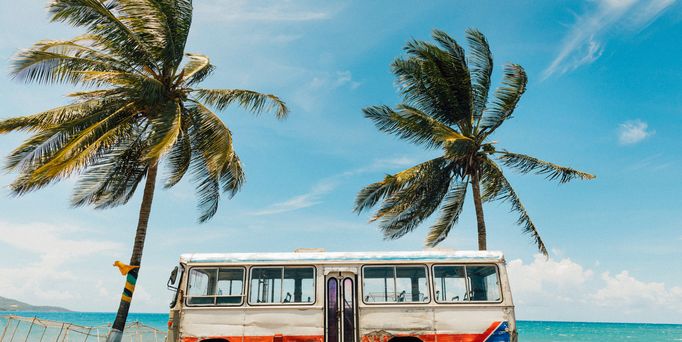 An einer Strandpromenade steht ein alter, rustikaler bunter Bus. Daneben und dahinter Kokospalmen.