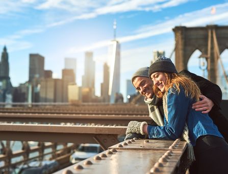 Ein junges Paar lehnt mit Winterkleidung am Geländer der Brooklynbridge. Im Hintergrund die Skyline von New York mit blauem Himmel.