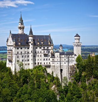 Ein märchenhaftes Schloss ragt in einer grünen, mitteleuropäischen Waldlandschaft empor und im Hintergrund befindet sich ein See.