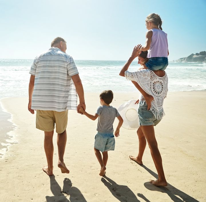 Eine Familie, bestehend aus einer Frau, 2 Kindern im Alter von ca. 4 bis 6 Jahren und einem älteren Mann, laufen an einem Sandstrand aufs Meer zu. Das Mädchen sitzt bei der Frau auf den Schultern und der Junge geht an der Hand des Mannes.