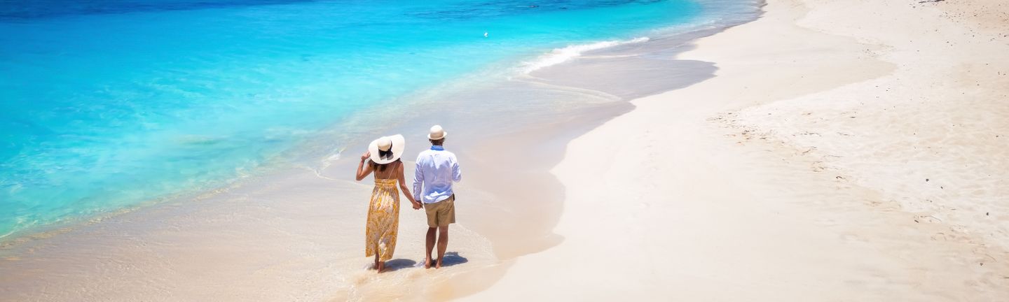 Ein Paar mit Sonnenhüten spaziert Hand-in-Hand an einem weißen, leeren Sandstrand entlang
