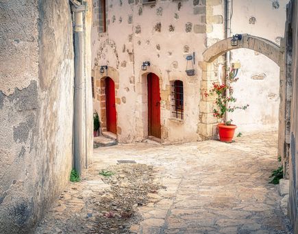 Eine griechische Gasse mit hellem Kopfsteinpflaster und einer Hausfront mit roten Türen.