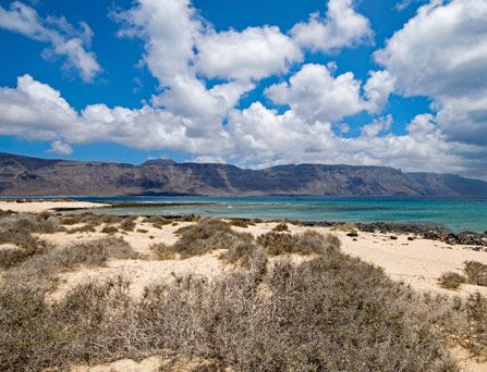 EIne karge, trockene Sandlandschaft vor dem Meer mit braunen, gigantischen Bergen im Hintergrund.