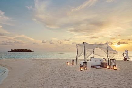 An einem weißen Strand steht ein Bett mit weißen Tüchern und Kerzen.