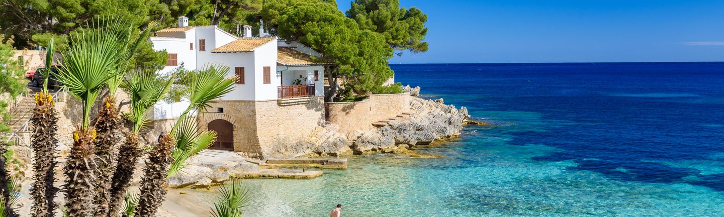 Eine wunderschöne, einsame Bucht mit türkis, blauem Wasser grenzt direkt an eine mallorquinische Finka mit Pinienbäumen und Palmen.