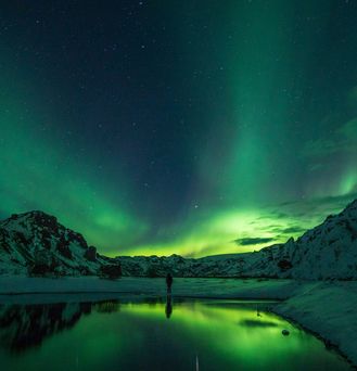 Der dunkel Nachthimmel wird von grünen Polarlichtern durchzogen. Eine Person steht, mit dem Rücken zugewandt und Wanderstöcken im weißen Schnee. Dahinter schneebedeckte Berge.
