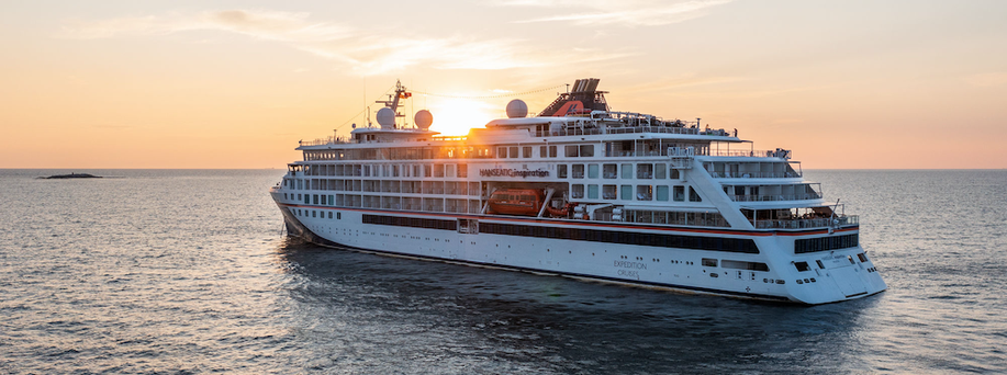 Ein mittelgroßes weißes Kreuzfahrtschiff auf dem offenen Meer. Die Sonne steht tief am Himmel und geht bald unter.