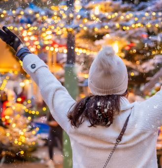 Eine Frau steht dem Betrachtenden mit dem Rücken zugewendet und armen in der Luft auf einem Weihnachtsmarkt. Sie trägt Mütze und Handschuhe und im Hintergrund sieht man viele gold-glitzernde Lichter.