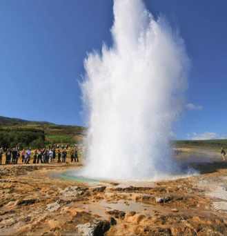 Aus dem Boden stößt explosionsartig eine riesige, kräftige, weiße Wasserfontäne nach oben in den blauen Himmel. Viele Menschen stehen darum herum und beobachten das Naturschauspiel.