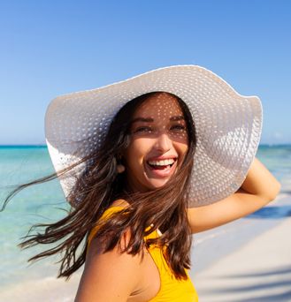 Eine Frau mit großem, weißen Sonnenhut läuft lachend an einem paradiesischen Strand entlang.