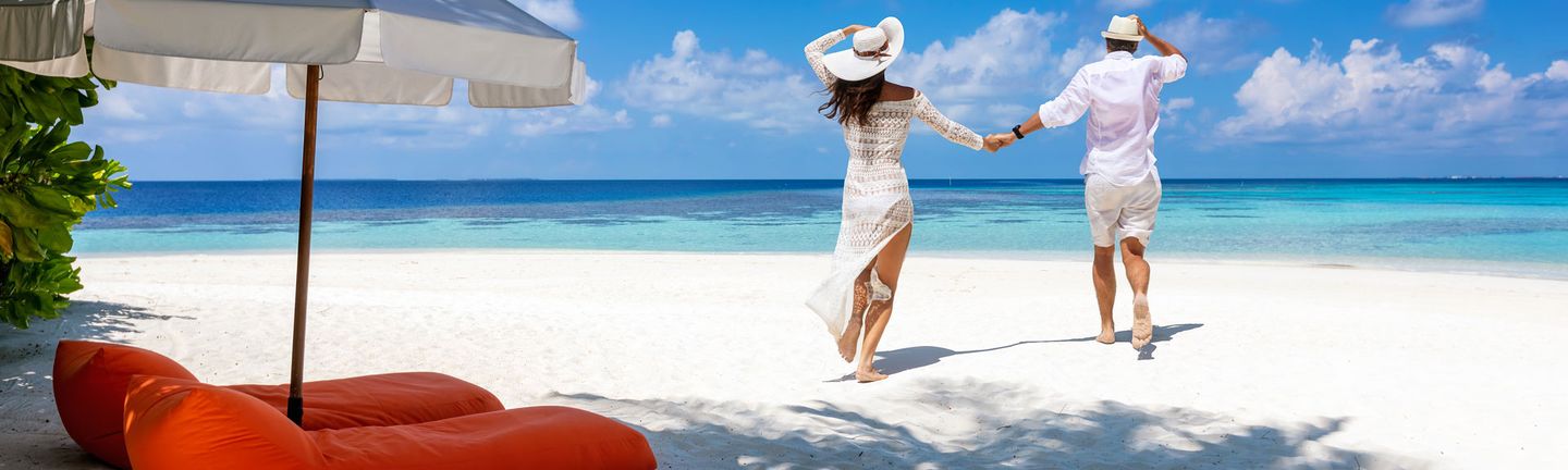 Ein Paar läuft Hand-in-Hand in sommerlicher, weißer Kleidung einen weißen Sandstrand auf das Meer zu