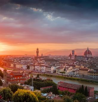 Die untergehende Sonne taucht Florenz  in einer violettes Abendlicht. Man sieht einen Fluss mit historischen Brücken und herausragende Kirchtürme und -uhren.