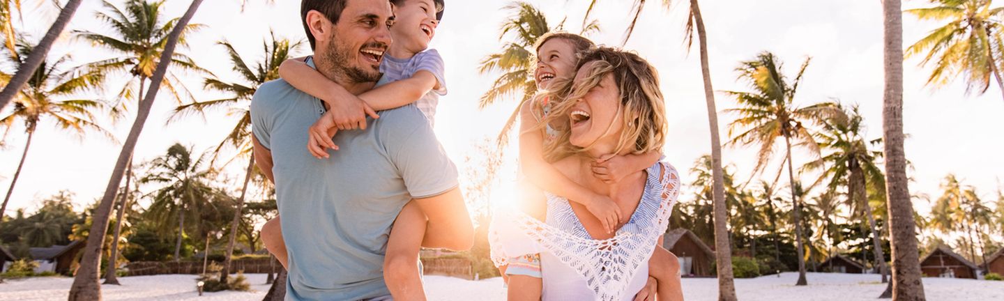 Eine junge vierköpfige Familie läuft lachend an einem Strand entlang. Der Vater hat den Sohn auf dem Rücken und die Mutter die Tochter. Alle tragen Sommerkleidung und lachen sich an.