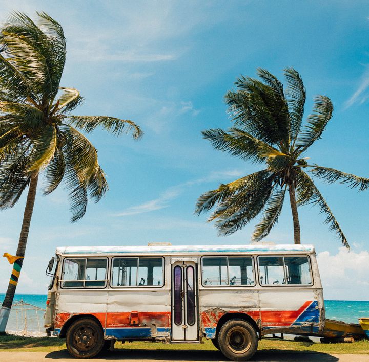 An einer Strandpromenade steht ein alter, rustikaler bunter Bus. Daneben und dahinter Kokospalmen.