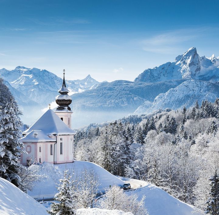 Eine kleine, weiß-rosa-farbene Kapelle ragt aus einer winterlichen Landschaft mit schneebedeckten Tannen und Berggipfeln hervor.