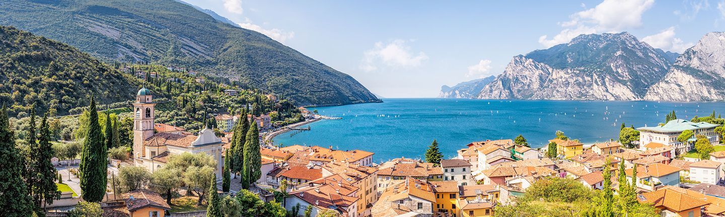 Ein mediterranes Dorf mit Kirche und orangenen Dächern liegt malerisch an einem tiefblauen See mit Segelbooten und einer beeindruckenden Bergkulisse.